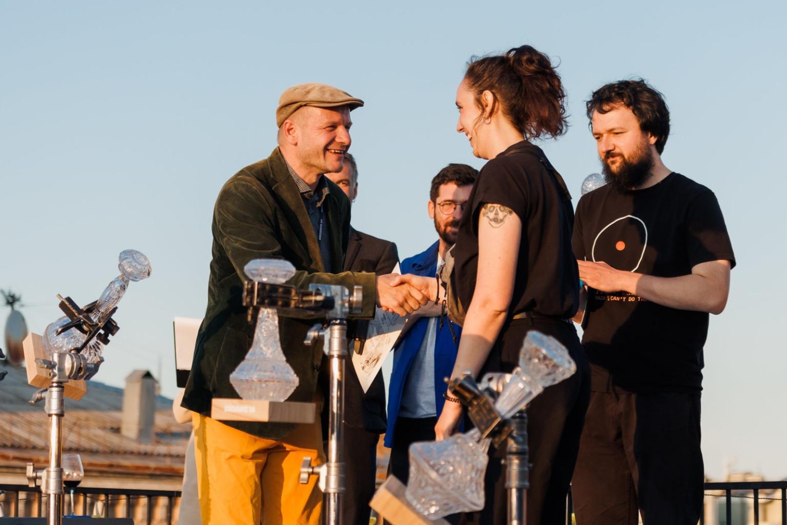 A small stage on a rooftop terrace. A young woman smiles and shakes the hand of a man who is presenting her with a certificate. Two other men stand in the background, also smiling.