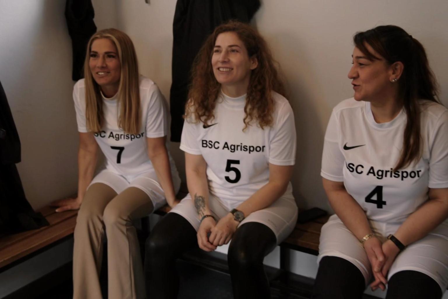 Drei Frauen in Fußballtrikots, auf denen BDC Agrisport un eine Nummer steht, sitzen auf einer Umkleidebank und grinsen in Richtung Kamera