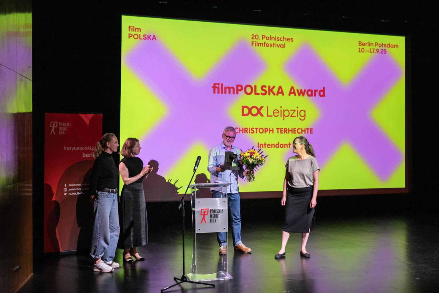 Preisverleihung auf einer Bühne: Vier Personen stehen vor einer großen Leinwand, eine Person hält Blumen und eine Urkunde, während die anderen applaudieren.