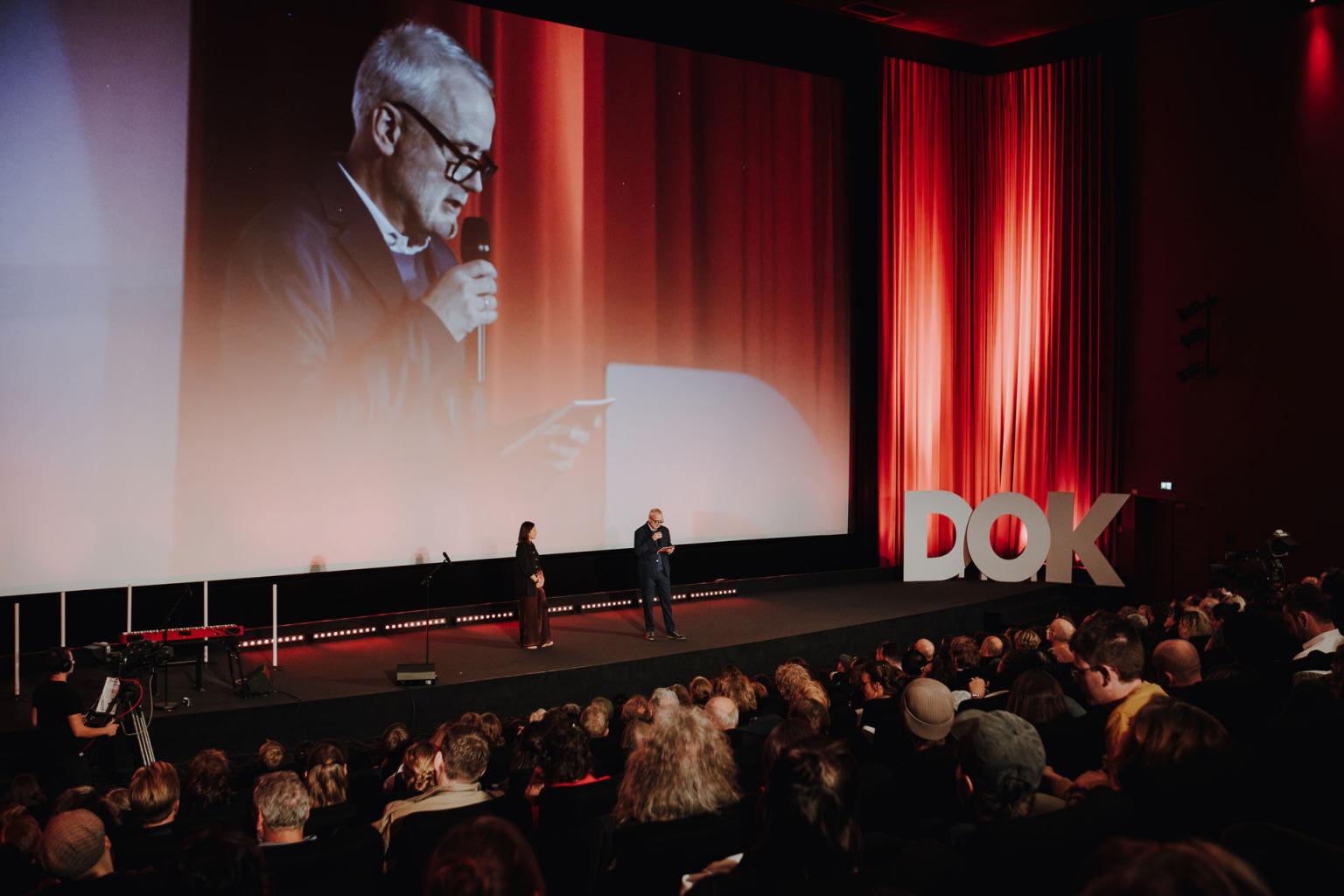 Two people stand on stage before a red curtain and large audience at DOK Leipzig; one speaks into a microphone, and the scene is projected on the big screen.
