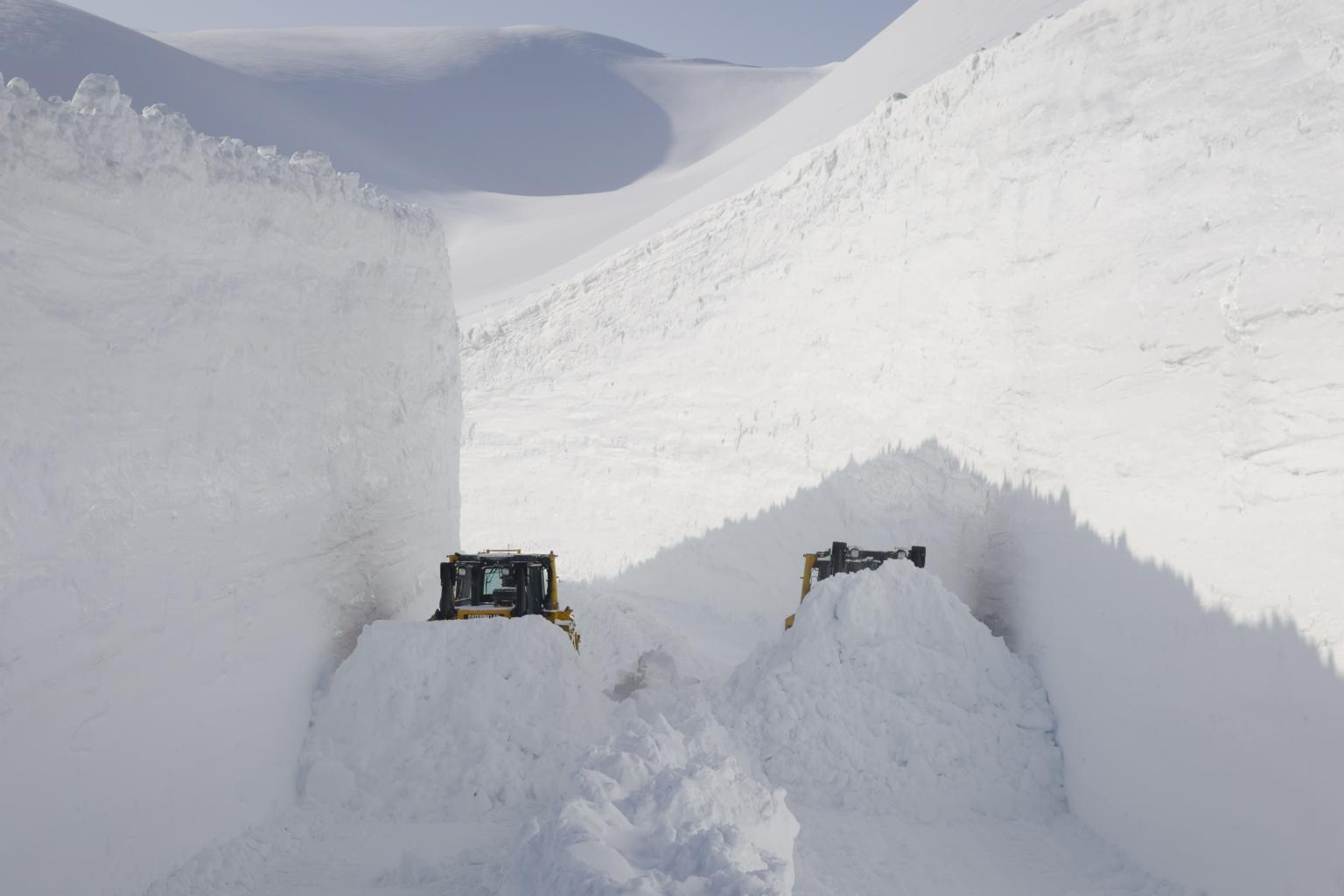 Two snowplows clear a deep, narrow path through towering snow in a snowy mountain landscape.