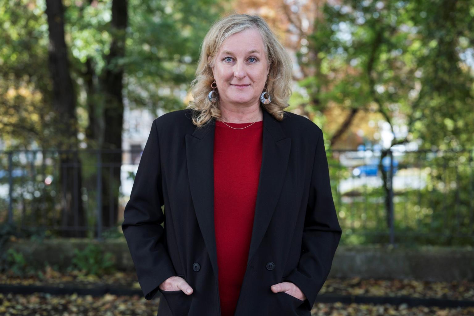A person with shoulder-length blonde hair stands outdoors in front of trees, wearing a black blazer over a red top and looking directly at the camera, gently smiling.