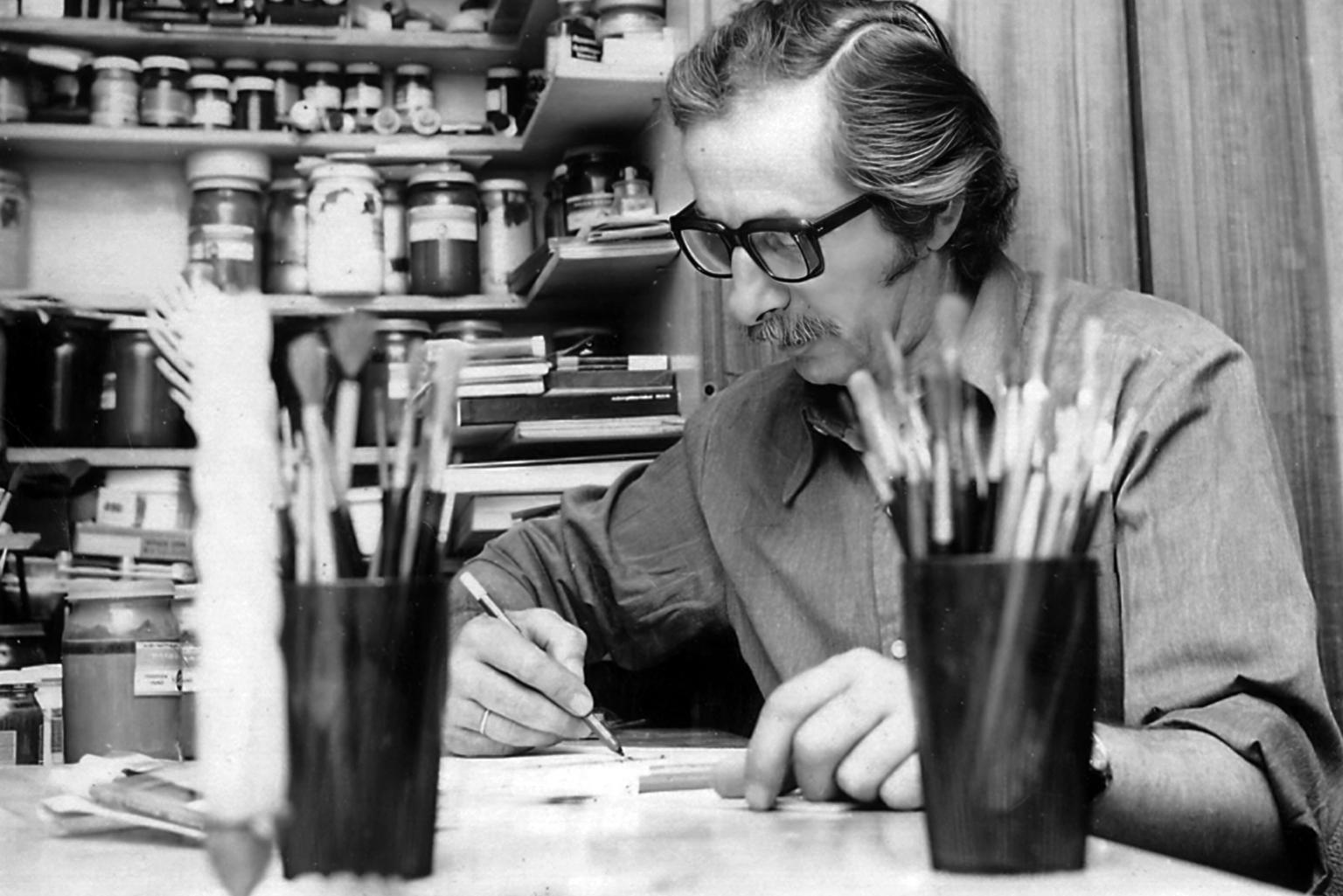 Black-white photo of a man sitting at a desk. He's sporting a huge mustache, thick dark glasses and combed back hair. In his hand he holds a pencil to a paper and he is surrounded by buckets of brushes and pens 