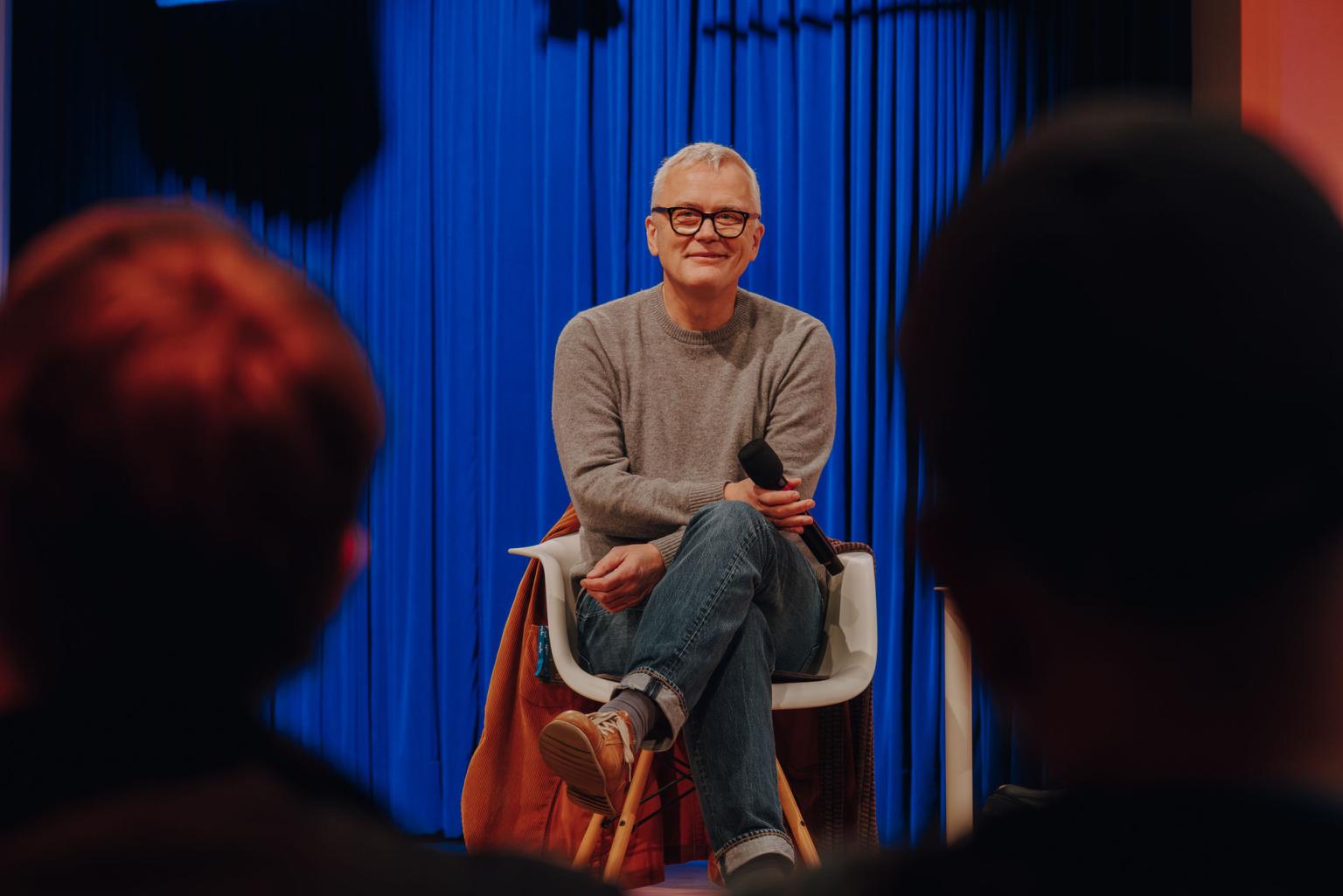 Mann mit grauen Haaren und Brille sitzt lächelnd mit Mikrofon vor blauem Vorhang, Publikum im Vordergrund.