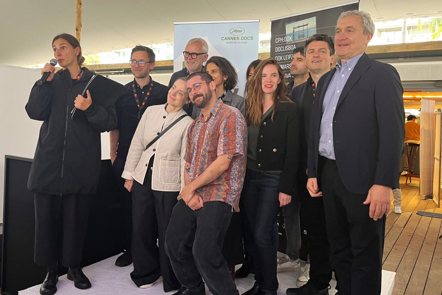 Gruppe von Filmfestival-Mitarbeitenden auf einer Bühne; links spricht eine Person ins Mikrofon, dahinter Banner internationaler Dokumentarfilm-Märkte.