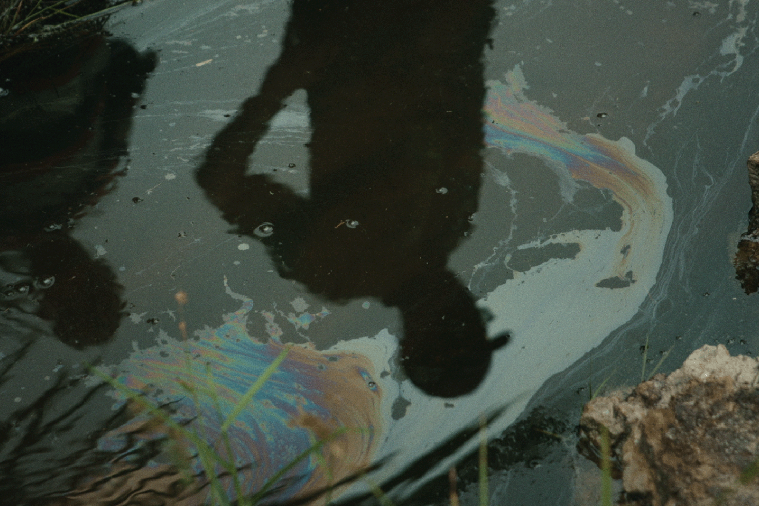 In ölig regenbogenfarbig schillerndem Wasser ist die Silhouette einer Person zu sehen, die den Kopf schwer neigt.
