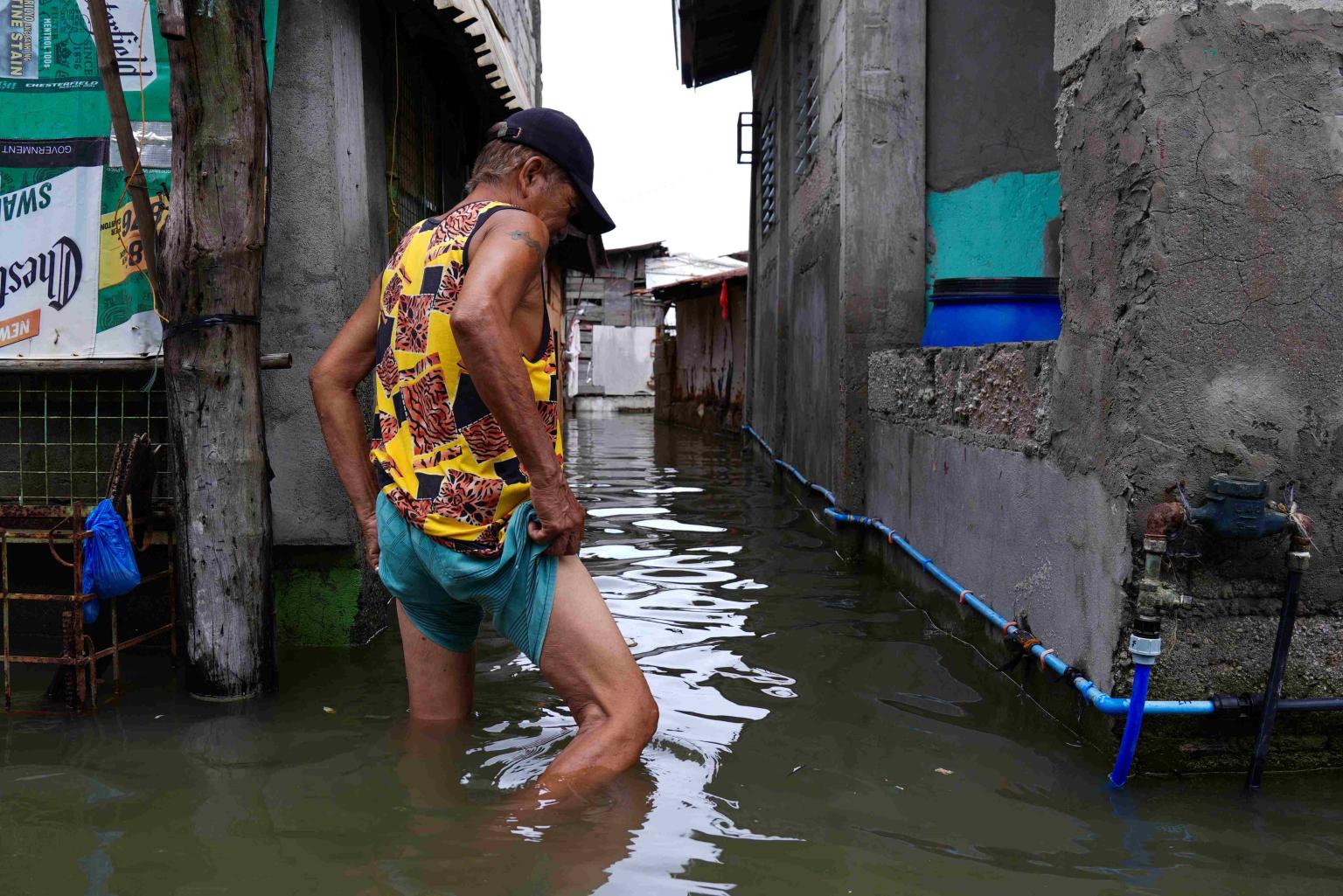 A man is wading through knee-deep water that has flooded a street between closely packed houses.