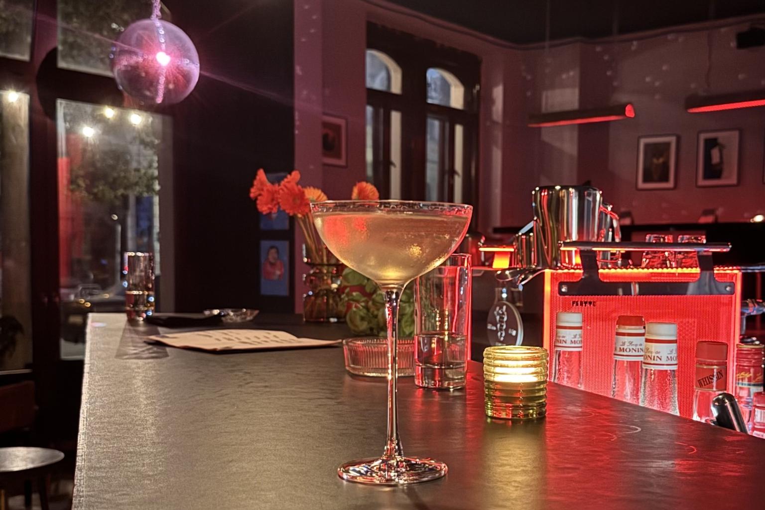 Eine abendliche leere Bar gemütlich bunt beleuchtet und mit Discokugel, im Vordergrund steht ein Cocktail in einem langstieligen Glass