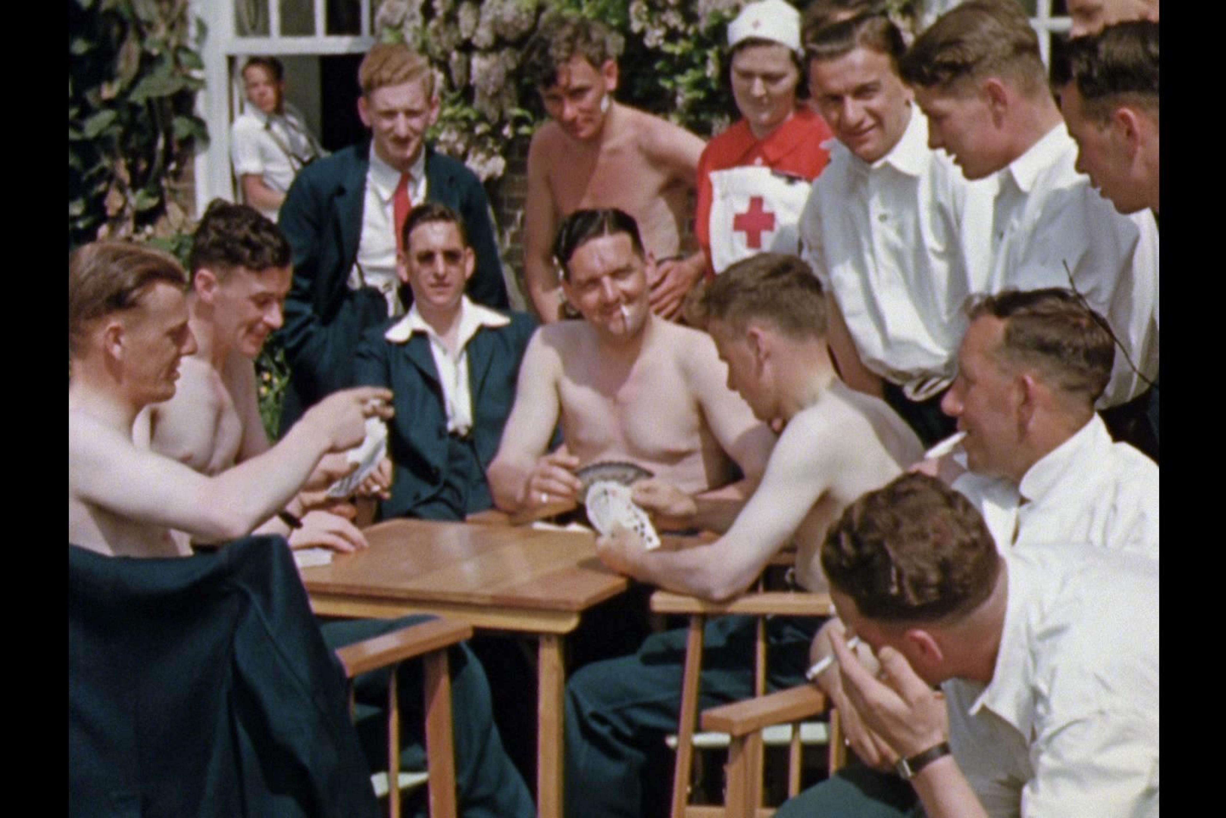 In a hospital garden: several bare-chested men are sitting around a table playing cards. Around them are several men and nurses watching.