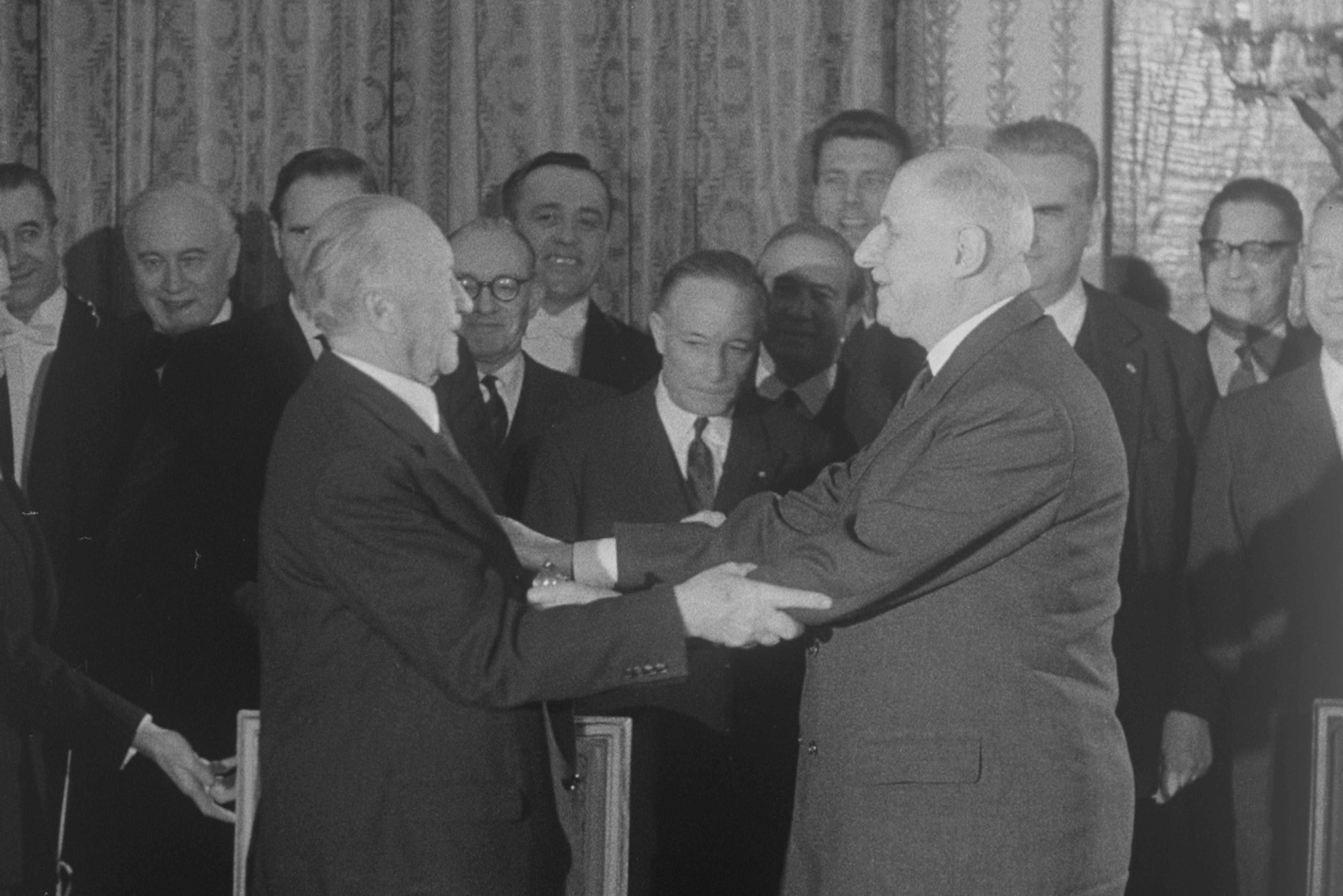 Black-and-white photo: German Chancellor Konrad Adenauer and French President Charles de Gaulle, both in suits, are shaking hands, smiling at each other. Behind them stand about 10 men watching them, all wearing suits.  
