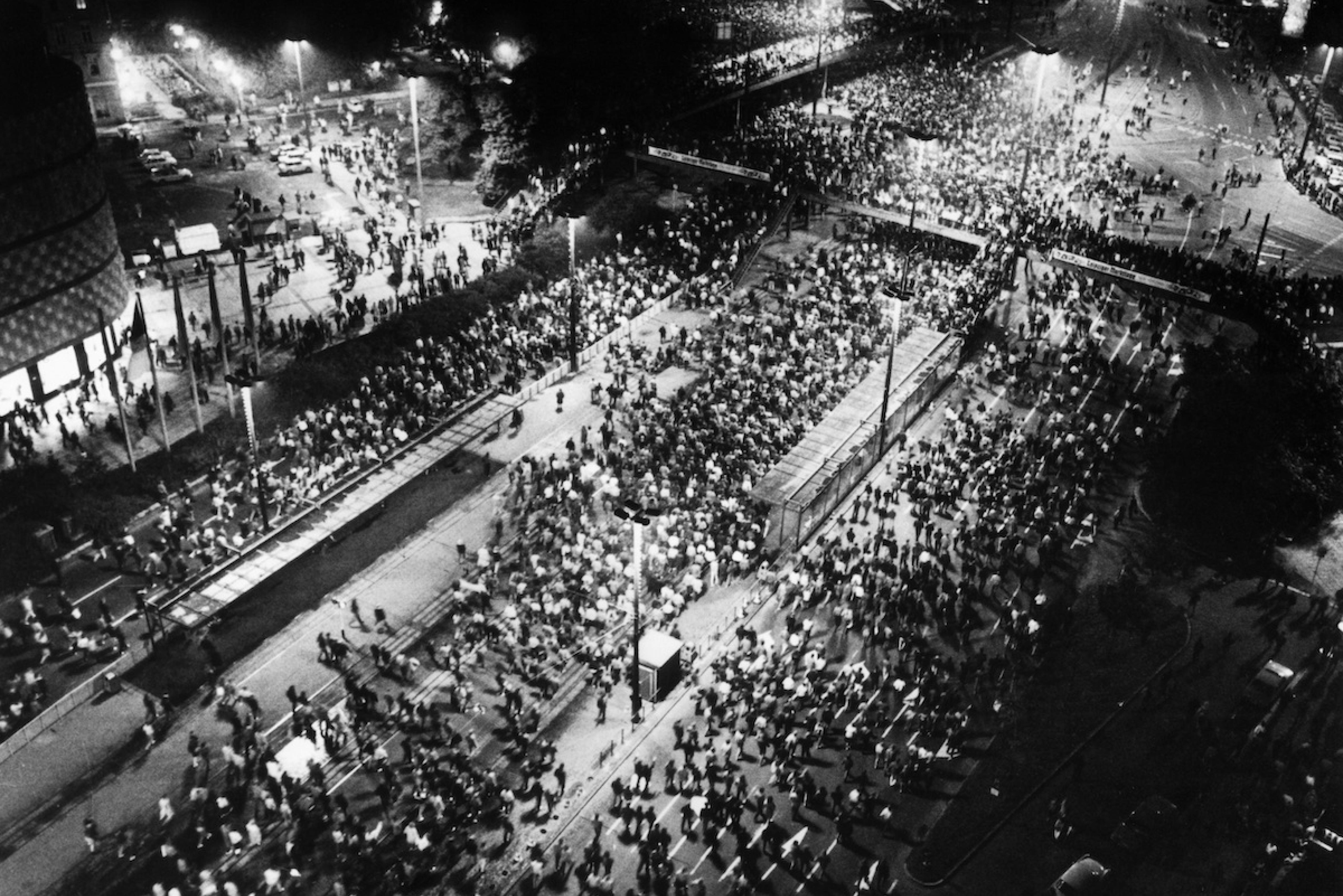 Black and white photo: A bird’s eye view of a big street and tram station in the evening. The street and station are crowded with people who are walking in a protest march.