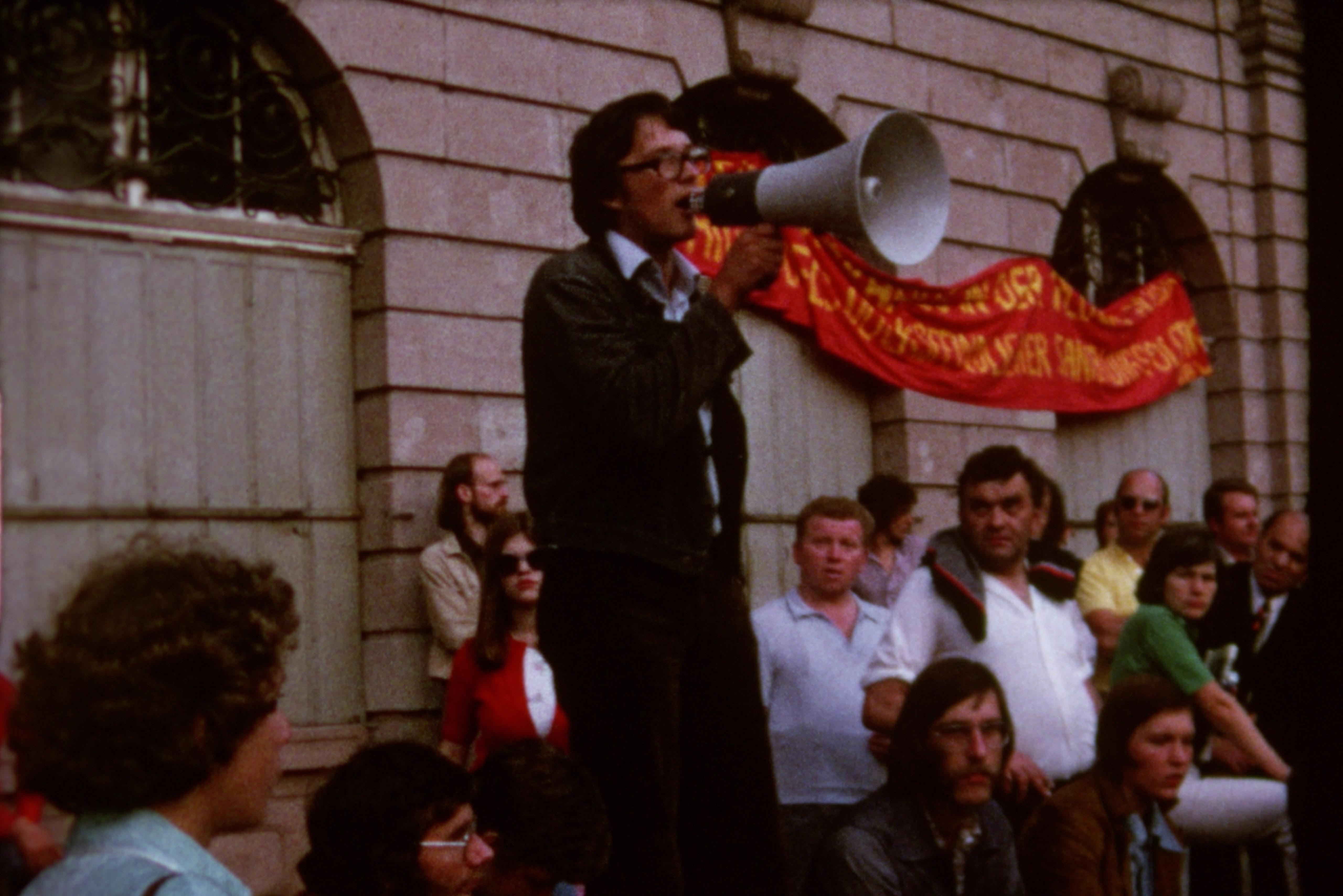 A man speaks into a megaphone. Around him, several people sit on the sidewalk in front of a building on which a red banner hangs.