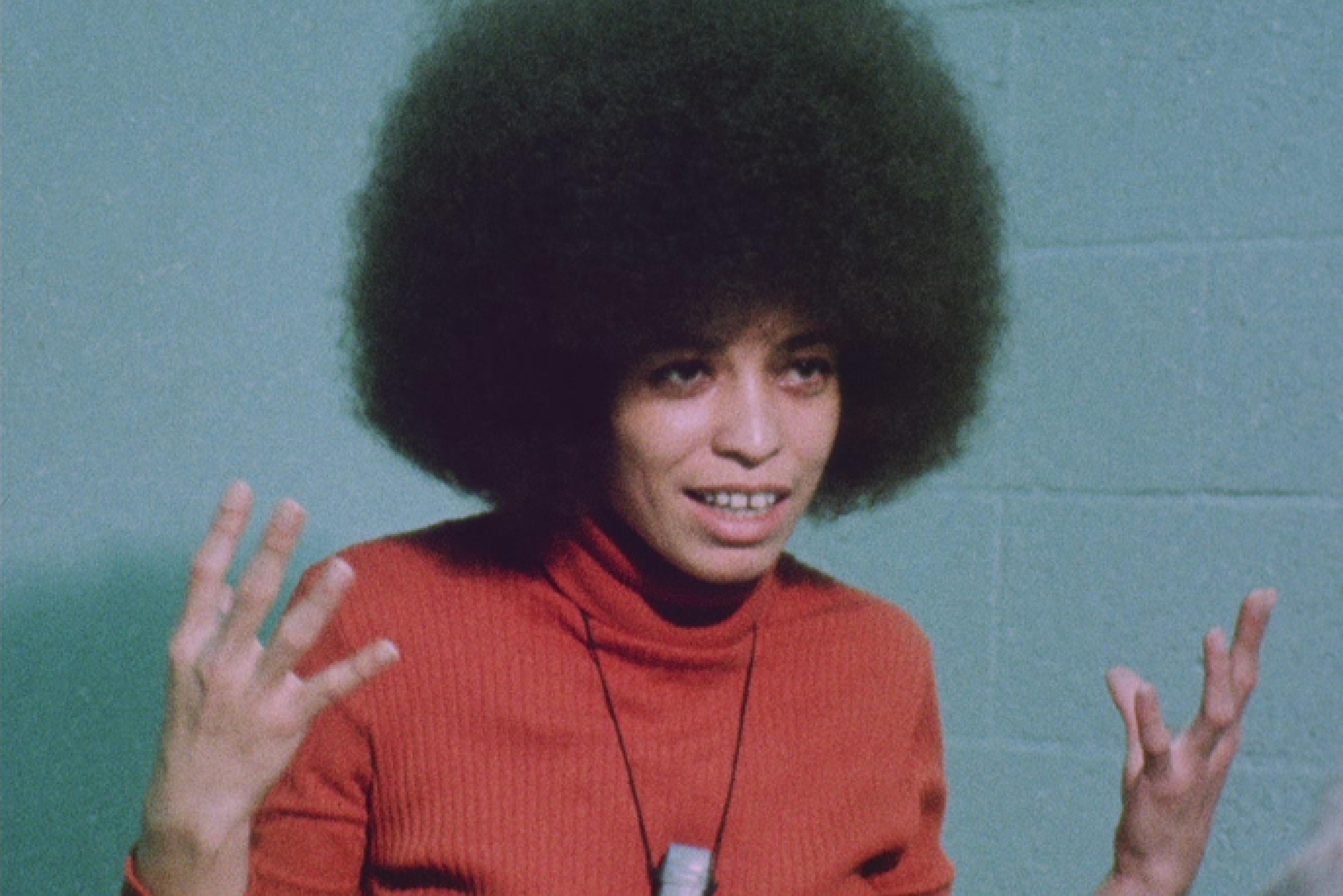 Portrait of Angela Davis. She is wearing an orange Turtleneck long sleeve and a microphone around her neck while gesturing with both hands.