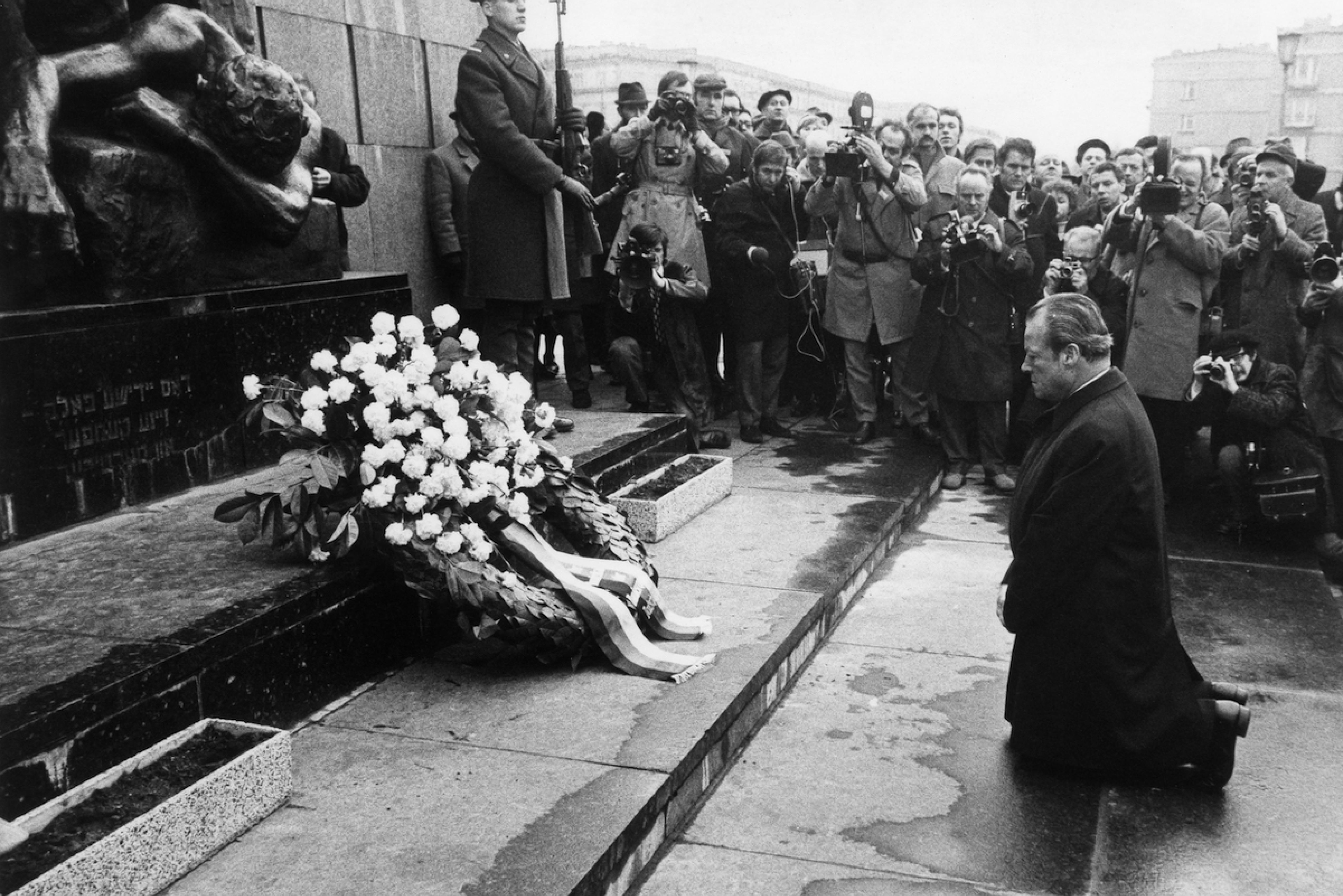 Black and white photo: German Chancellor Willy Brandt kneeling in front of the memorial to the Warsaw Ghetto. In the background a group of journalists and photographers, and one soldier.