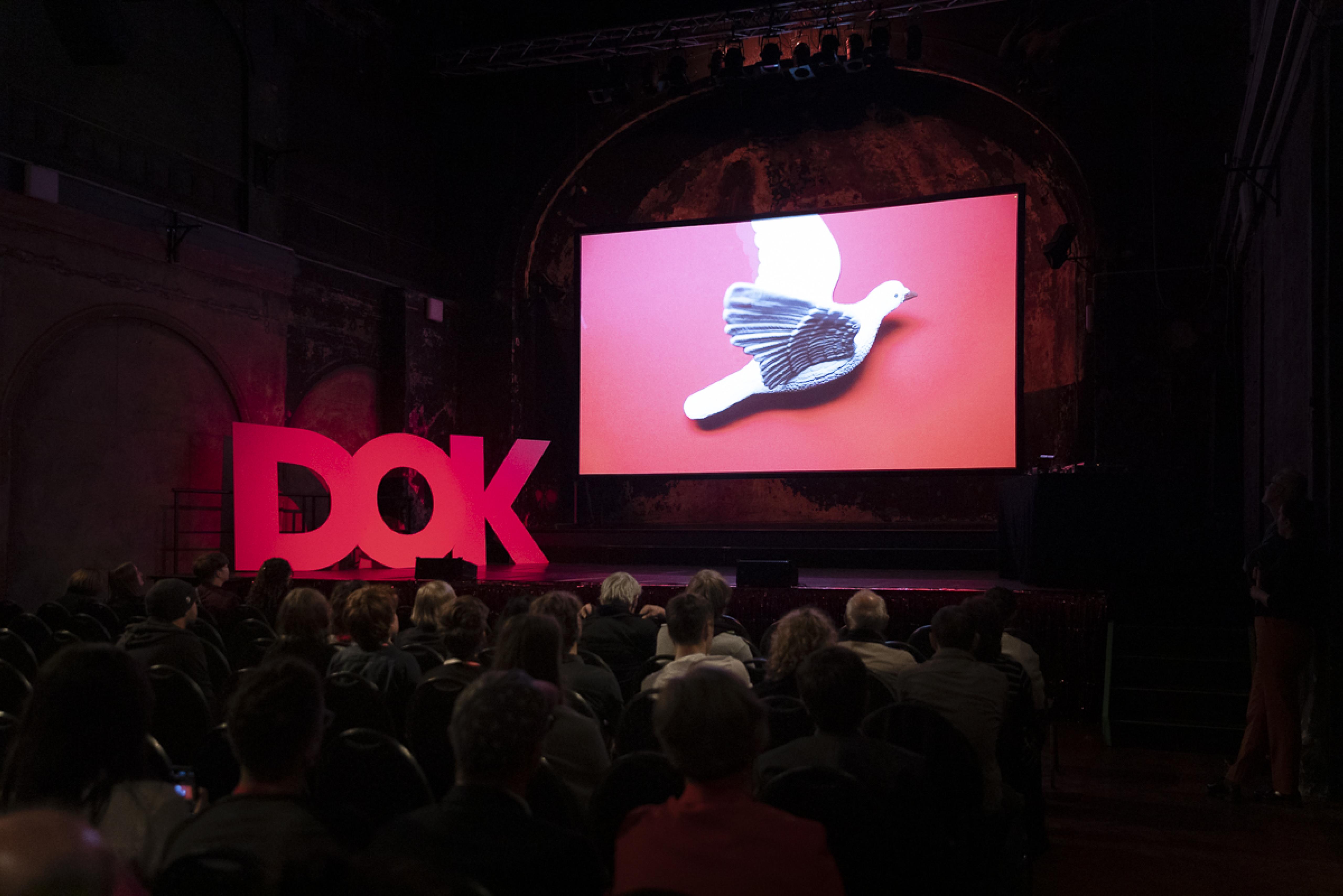 A stage in a big hall. On stage three huge letters "D-O-K" and a big screen showing a white dove.