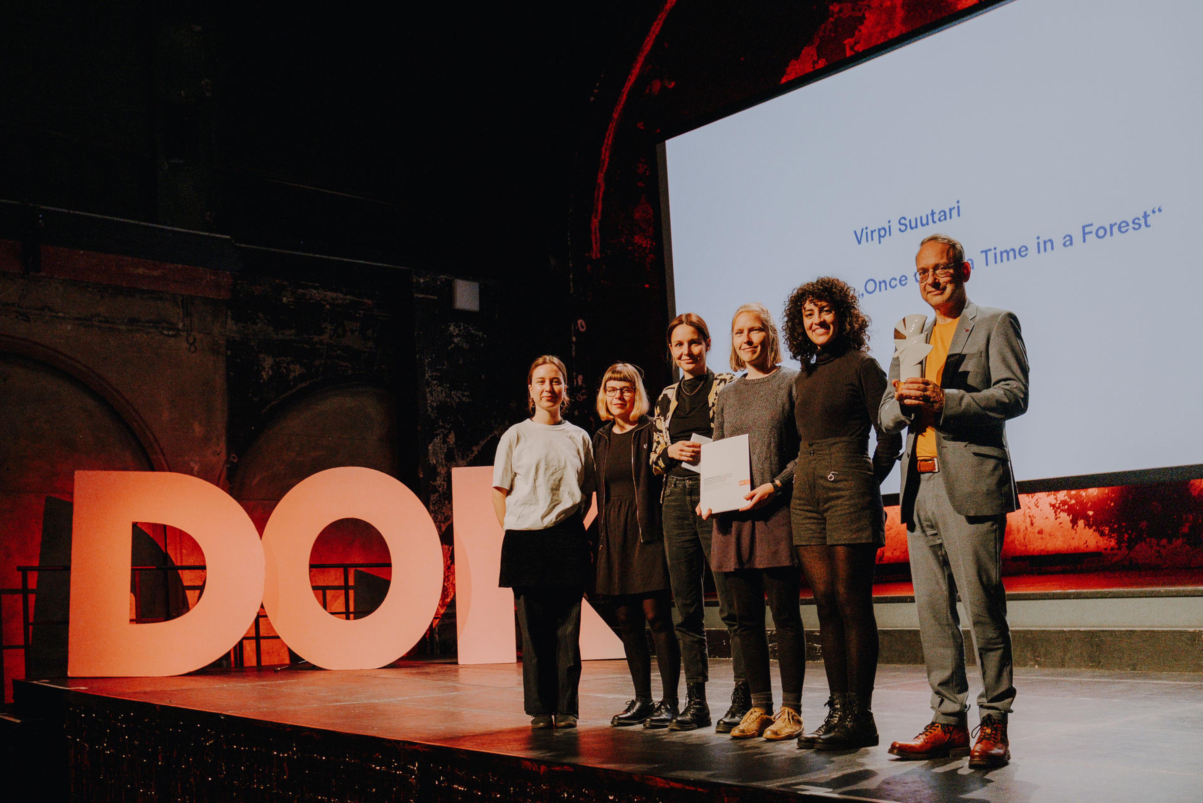 Die Publikumsjury auf der Bühne der Dok Leipzig Preisverleihung mit dem Preisträger, der die Goldene Taube-Statue in der Hand hält