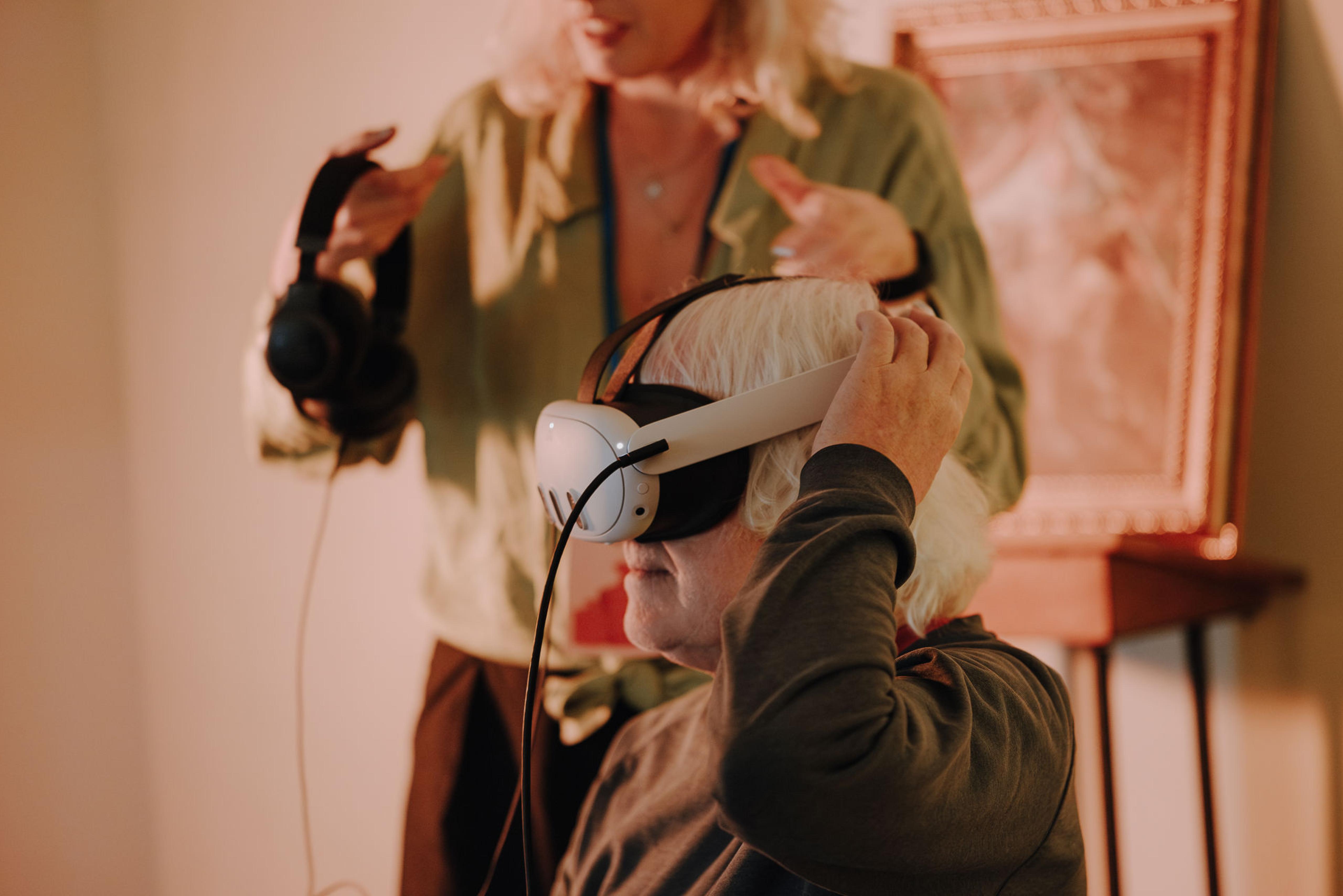 A person with white hair is pulling vr glasses over his head, behind him one of the project teams is offers him headphones