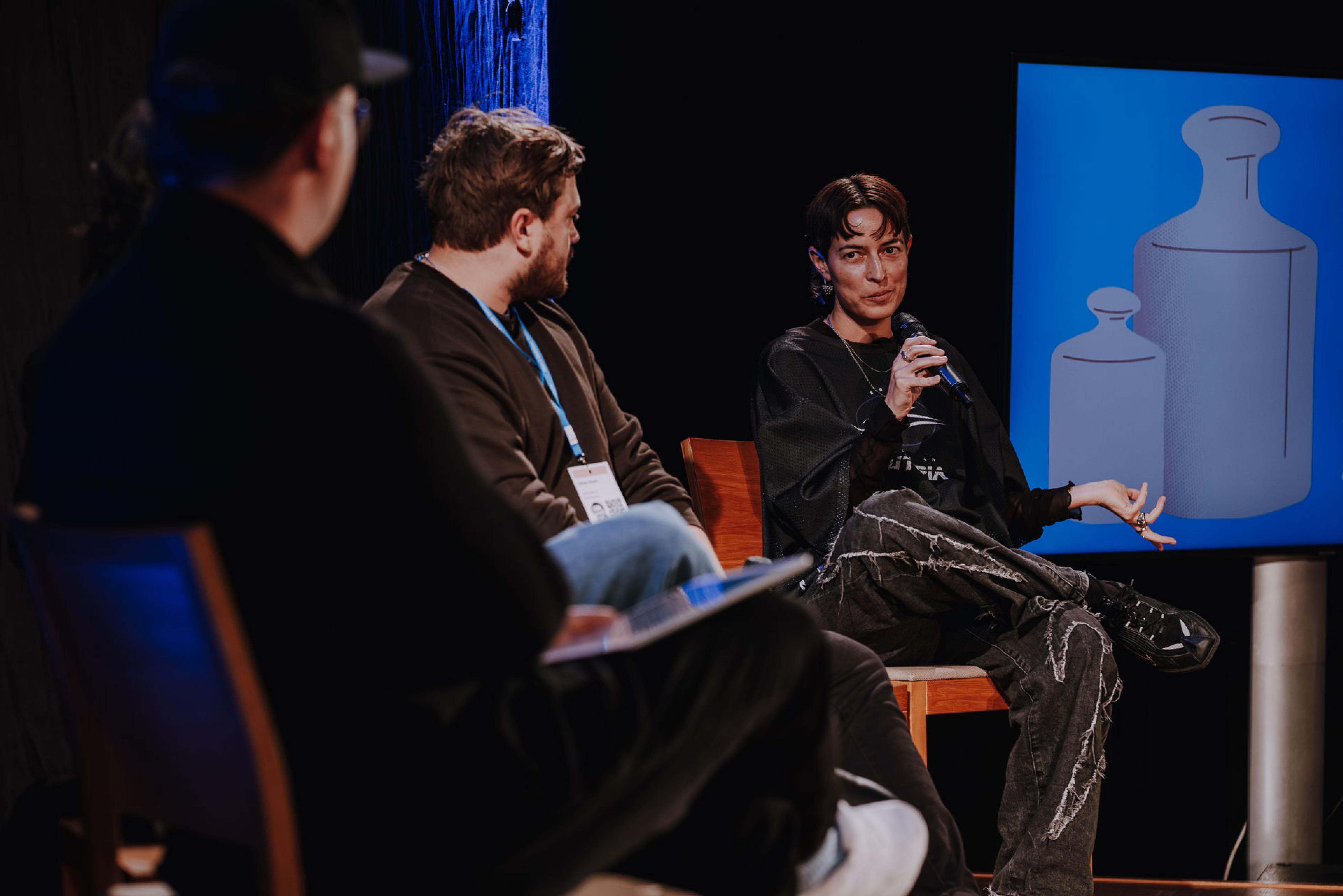 A panel with three people on stage, one person talks into a microphone and gesticulates