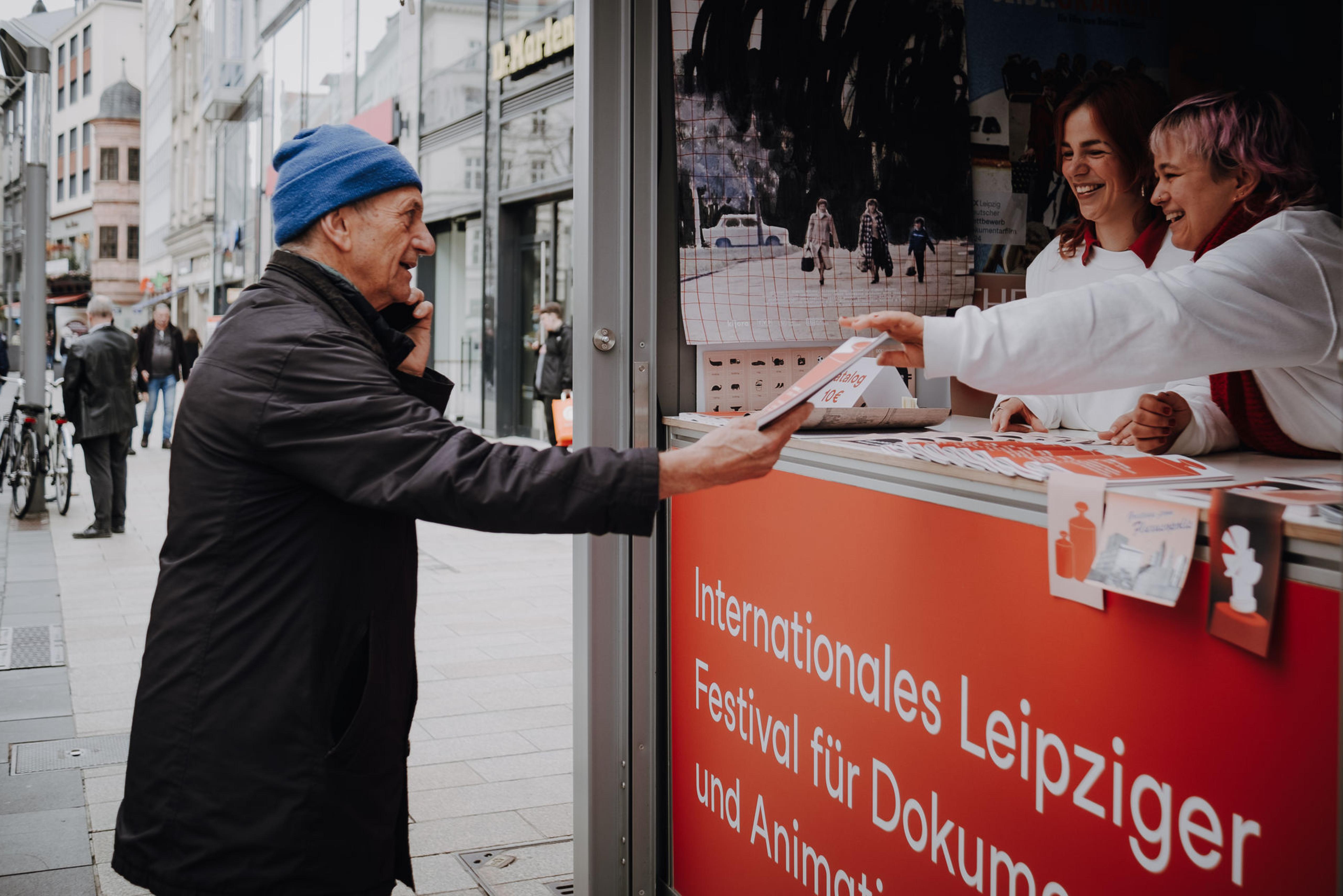 Zwei Helferinnen in der DOK Box reichen einem Mann das DOK Programmheft