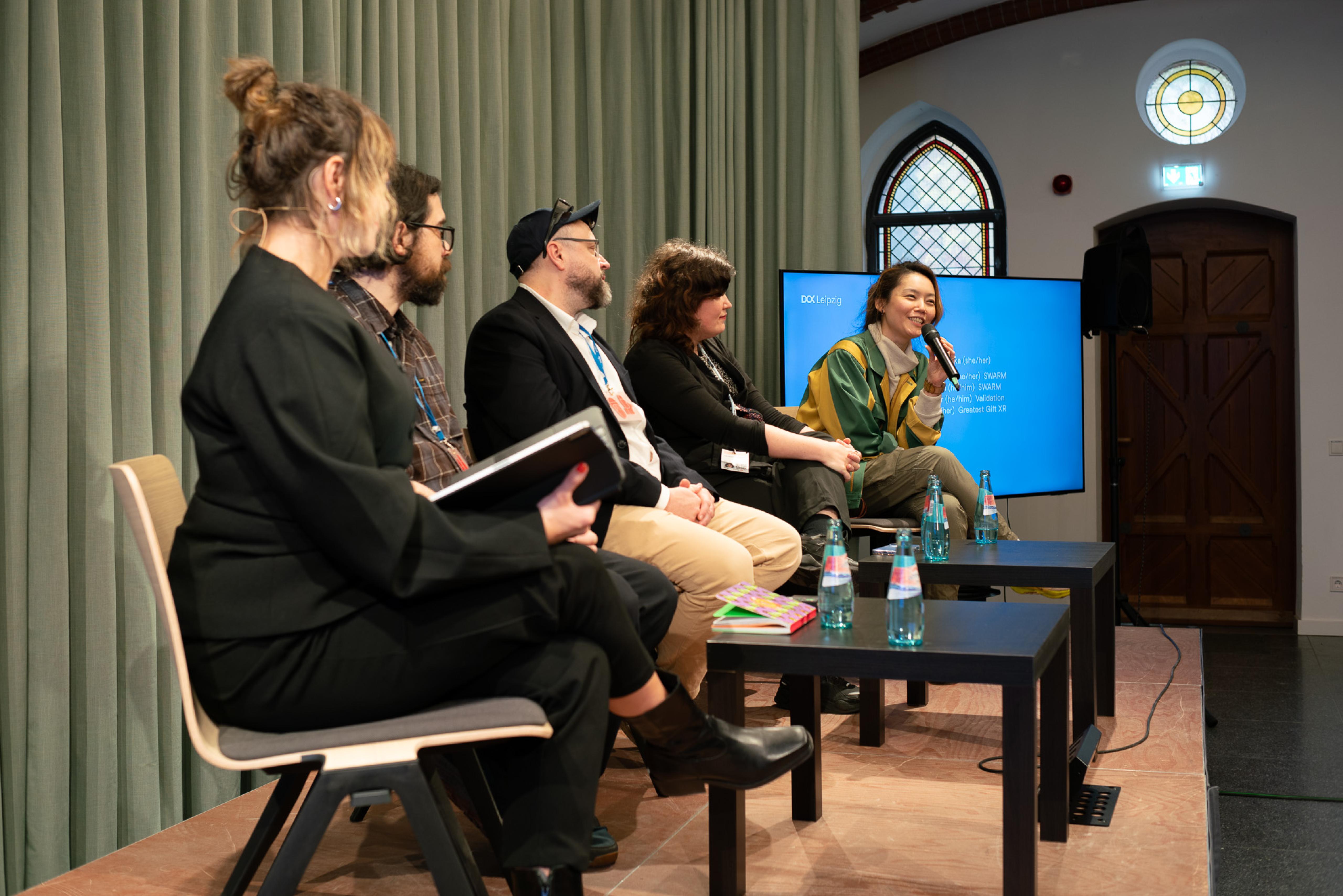 On a small stage, five people sit in a row, four of them looking at a woman speaking into a microphone