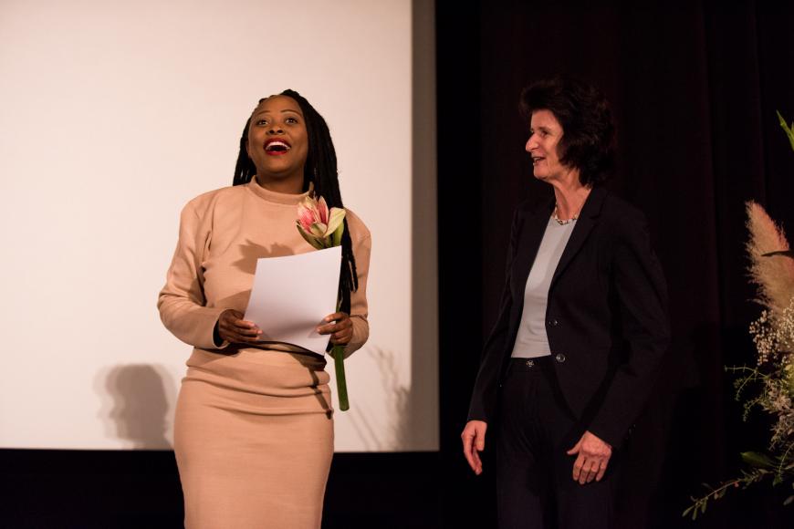 Two women on a stage: the black woman on the left side is the director of the awarded film "Black women and sex" and is holding a certificate and flowers. The woman standing next to her is the state minister for art and culture. 