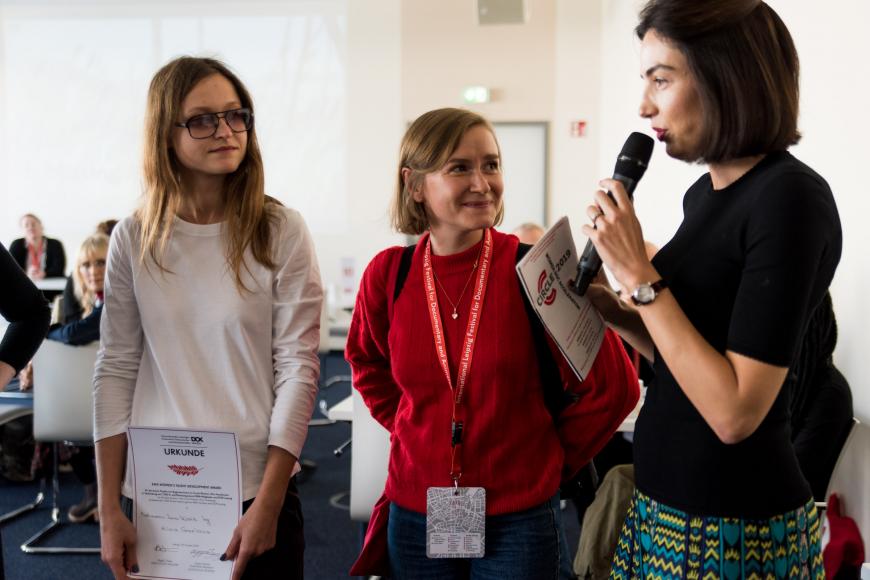 Two women are looking at a woman standing next to them and speaking into a microphone.