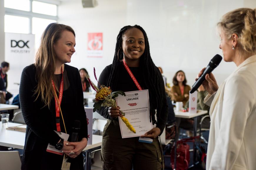 Three women standing side by side, the woman in the middle holds a certificate in her hands and is smiling into the camera.