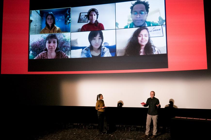 A woman and a man moderating in front of a cinema screen, where you can see the faces of six persons.