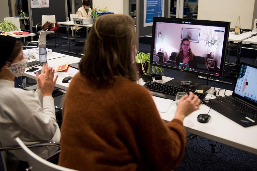 Ulrike Schmidt sitzt vor einem Computerbildschirm, darin ist das Video von einer Frau in ihrem Wohnzimmer zusehen, die lächelt und sich mit der Hand auf ihr Herz fasst 