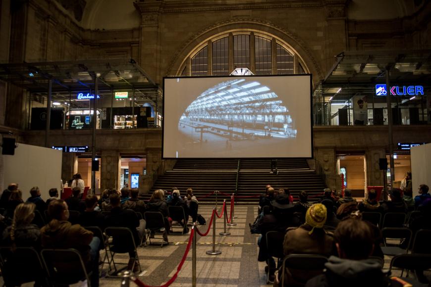 A screening in the big hall of the Leipzig main station, a big audience is watching a train on the screen