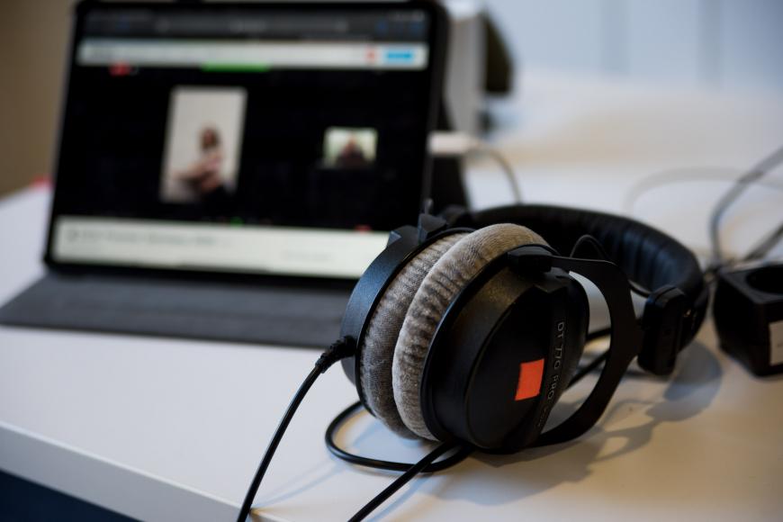 Ein großer Kopfhörer mit Kabel liegt auf einem Schreibtisch, im Hintergrund sind unscharf die Umrisse eines Laptops zu sehen.