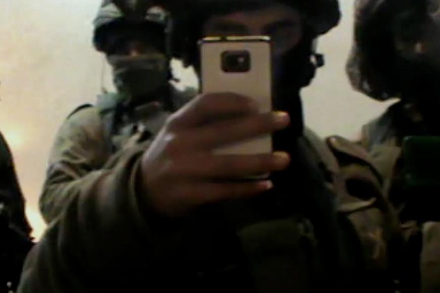 Filmstill: Vier Soldaten in Uniform und mit Helmen stehen nebeneinander, einer hält ein Smartphone hoch und macht ein Foto.