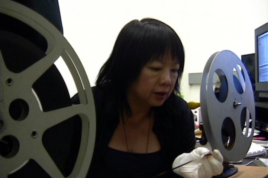 Mary Stephen sitzt an einem analogen Schneidetisch zwischen zwei Filmrollen. Sie blickt auf den Film, den sie in ihrer Hand hält.