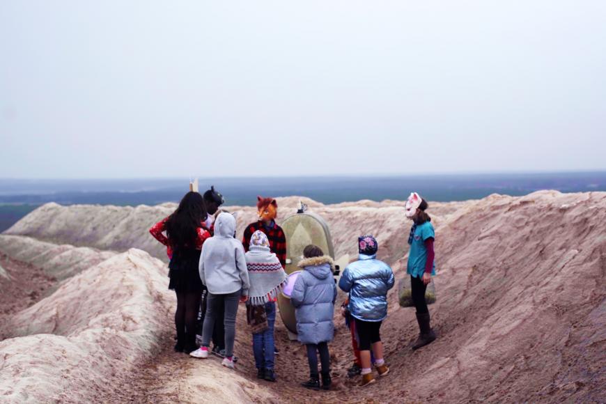 Eine Gruppe von acht Kindern, die Tiermasken tragen, stehen in einer hügeligen Steinlandschaft im Kreis vor einer menschengroßen Kiste, im Hintergrund das Meer