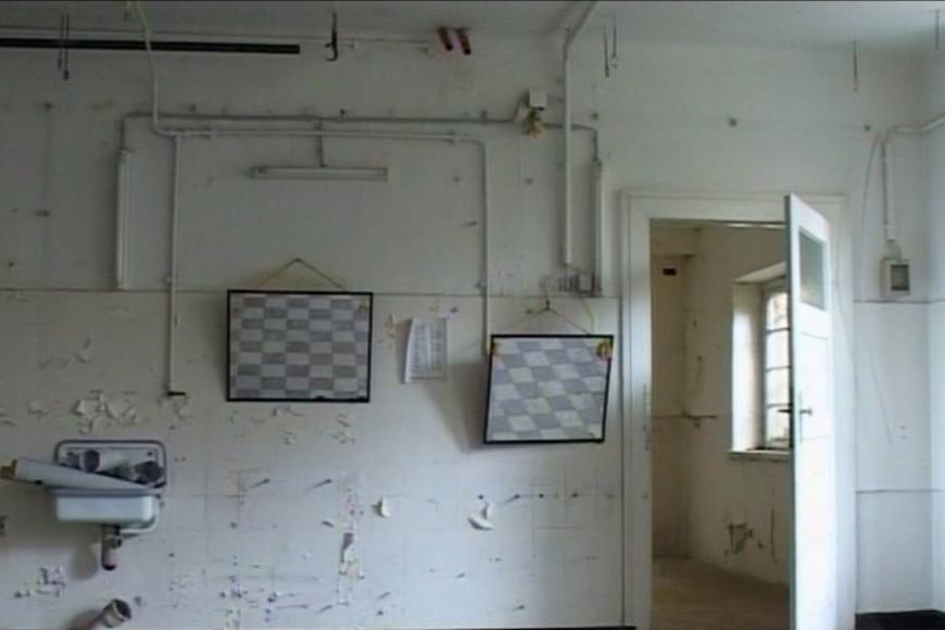Filmstill: Innenansicht eines leerstehenden Gebäudes, Tapete blättert von den kahlen Wänden, zwei Spiegel und ein kleines Waschbecken sind noch an der Wand montiert.