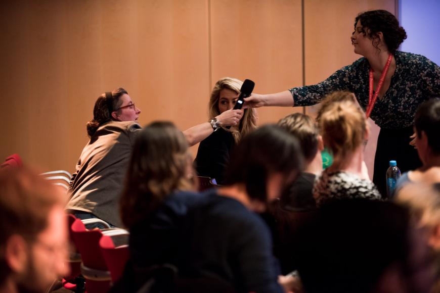 Besucher*innen eines Talks bei DOK Leipzig. Eine Mitarbeiterin reicht ein Handmikrofon an eine Zuschauerin weiter.