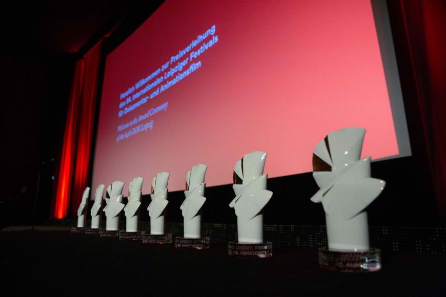 Acht Preisstatuetten der Goldenen und Silbernen Tauben stehen aufgereiht nebeneinander auf einer Bühne. Hinter ihnen eine große Leinwand.