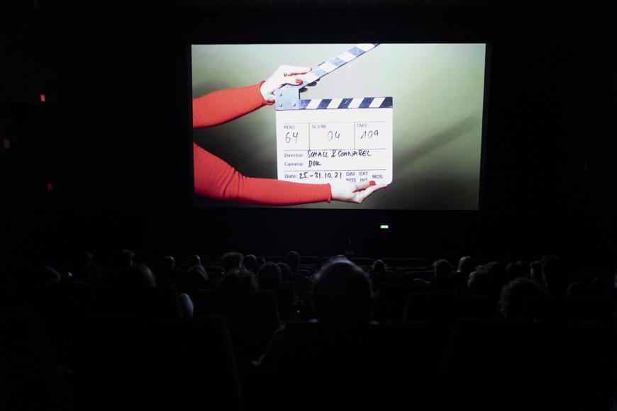 Blick über die Sitzreihen in einem dunklen Kinosaal auf die erleuchtete Leinwand. Darauf wird gerade eine Filmklappe von zwei Händen zugeschlagen.