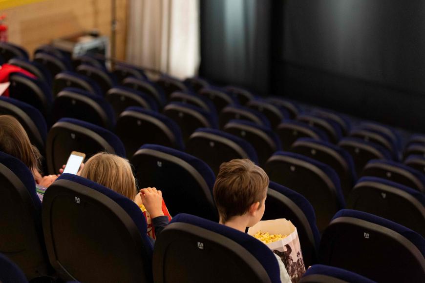Im Kino: Drei Kinder sitzen mit Popcorn-Bechern auf ihren Sitzplätzen in einer der vorderen Reihen. Sie warten darauf, dass der Film beginnt.