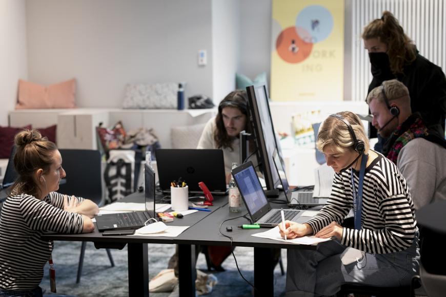 Fünf Personen gruppieren sich sitzend und stehend um einen großen quadratischen Arbeitstisch mit mehreren Laptops. Eine Frau (rechts im Bild) macht sich  Notizen auf einen DIN A4-Zettel.