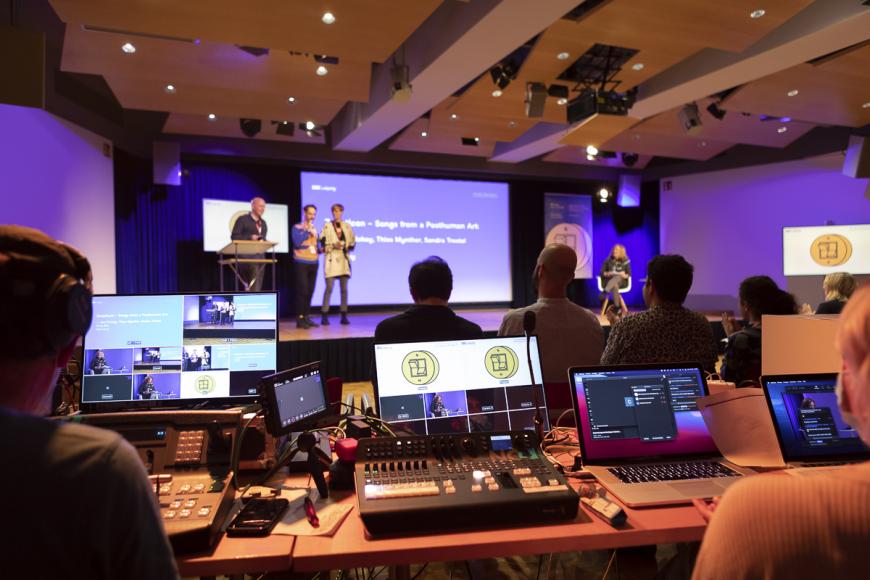 A presentation at DOK Industry: In the foreground a huge live editing unit with four displays and a control desk. In the background, three persons on stage, equipped with microphones.