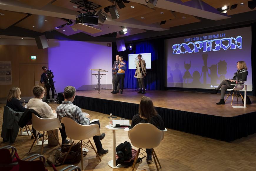 A pitch at DOK Industry: Three persons present on stage in front of a four-person panel. Moderator Elisabeth Klinck  sits at the left side of the stage while listening.