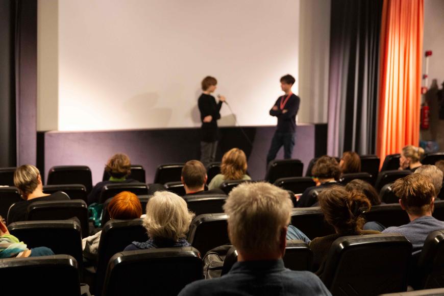 Blick über gut gefüllte Zuschauerreihen in einem Kinosaal in Richtung Leinwand. Das Licht im Saal ist an, vor der Leinwand stehen zwei Personen einander zugewandt. Die Person links spricht in ein Handmikro.