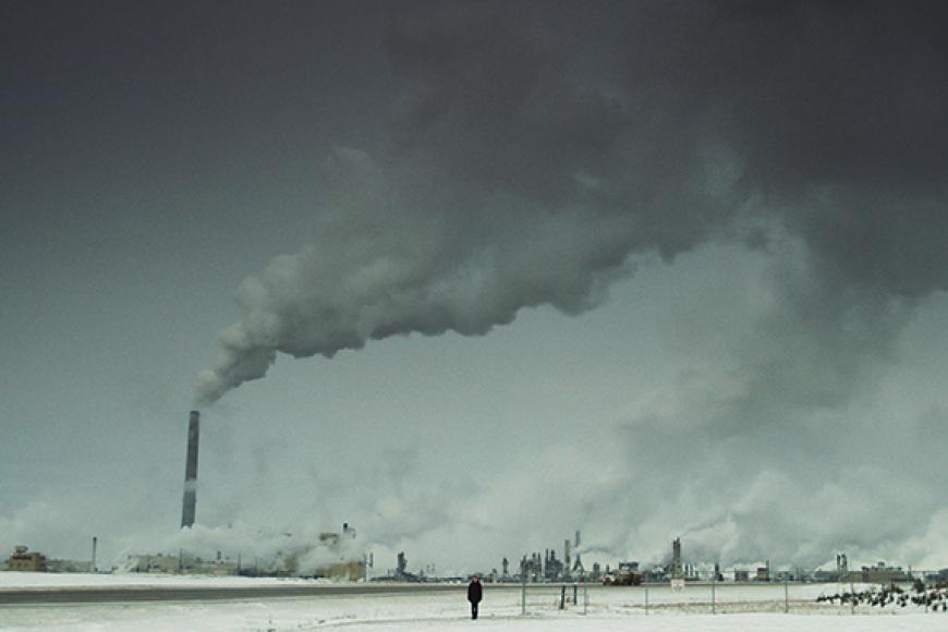 Eine wüstenähnliche, karge Ebene. Am Horizont sind viele rauchende Schornsteine mehrerer Raffinerien zu sehen. Mitten in dieser Szenerie steht eine schwarz gekleidete Person. 