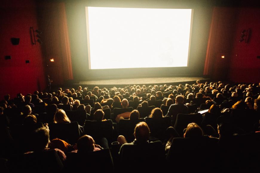 Ein vollbesetztes Kino bei einer Filmvorführung. Die Leinwand strahlt helles, weißes Licht über die Köpfe der Zuschauer*innen.