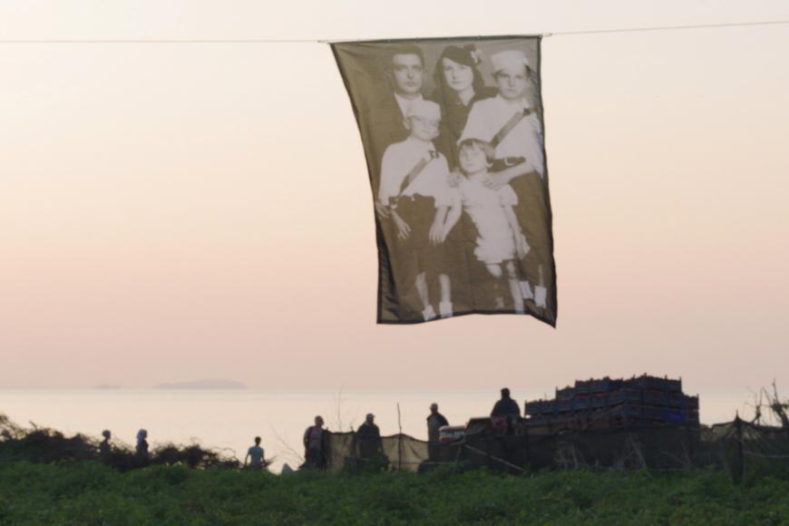 Ein bedrucktes Tuch hängt an einer Wäscheleine, auf dem Tuch ist das schwarz-weiß Foto einer Familie mit zwei Kindern. 