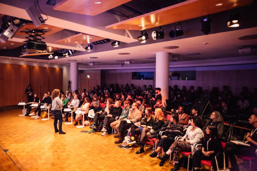 A woman stand in front of an audience in a conference room. She speaks into a microphone.