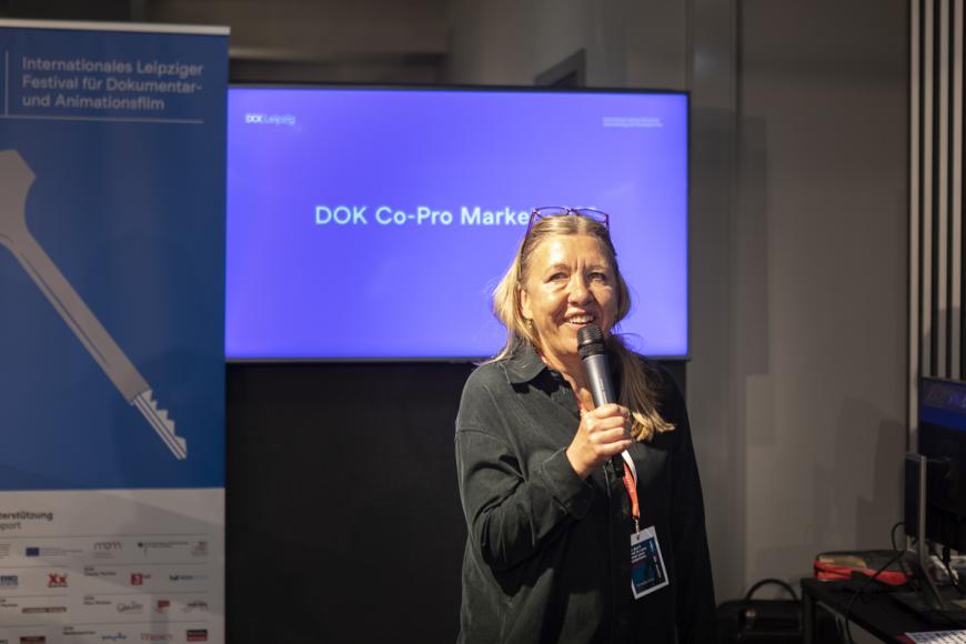 Nadja Tennstedt, head of DOK Industry, stands in front of a screen showing the text: "DOK Co-Pro Market". She smiles while speaking into a hand microphone.