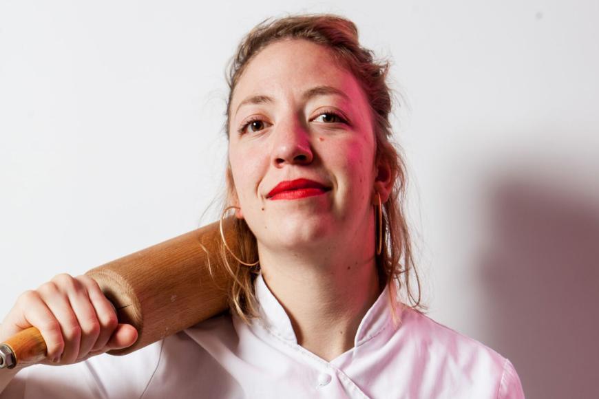 Portrait einer Frau, die einen Kochkittel trägt. Sie hat ein Nudelholz lässig auf ihre Schulter gelegt und schaut leicht lächelnd direkt in die Kamera.