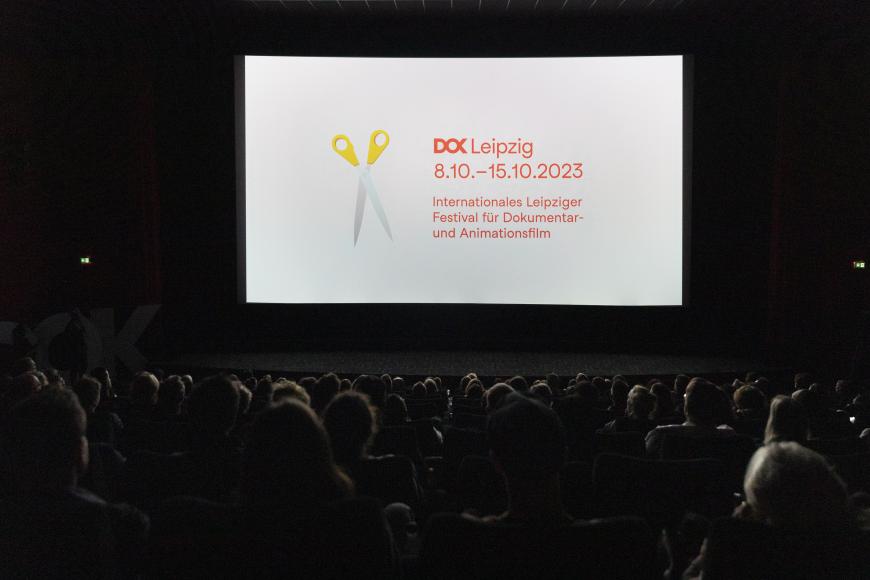 Ein voller Kinosaal bei einer Filmvorführung. auf der Leinwand ist das Festivalmotiv mit dem Logo von DOK Leipzig zu sehen.